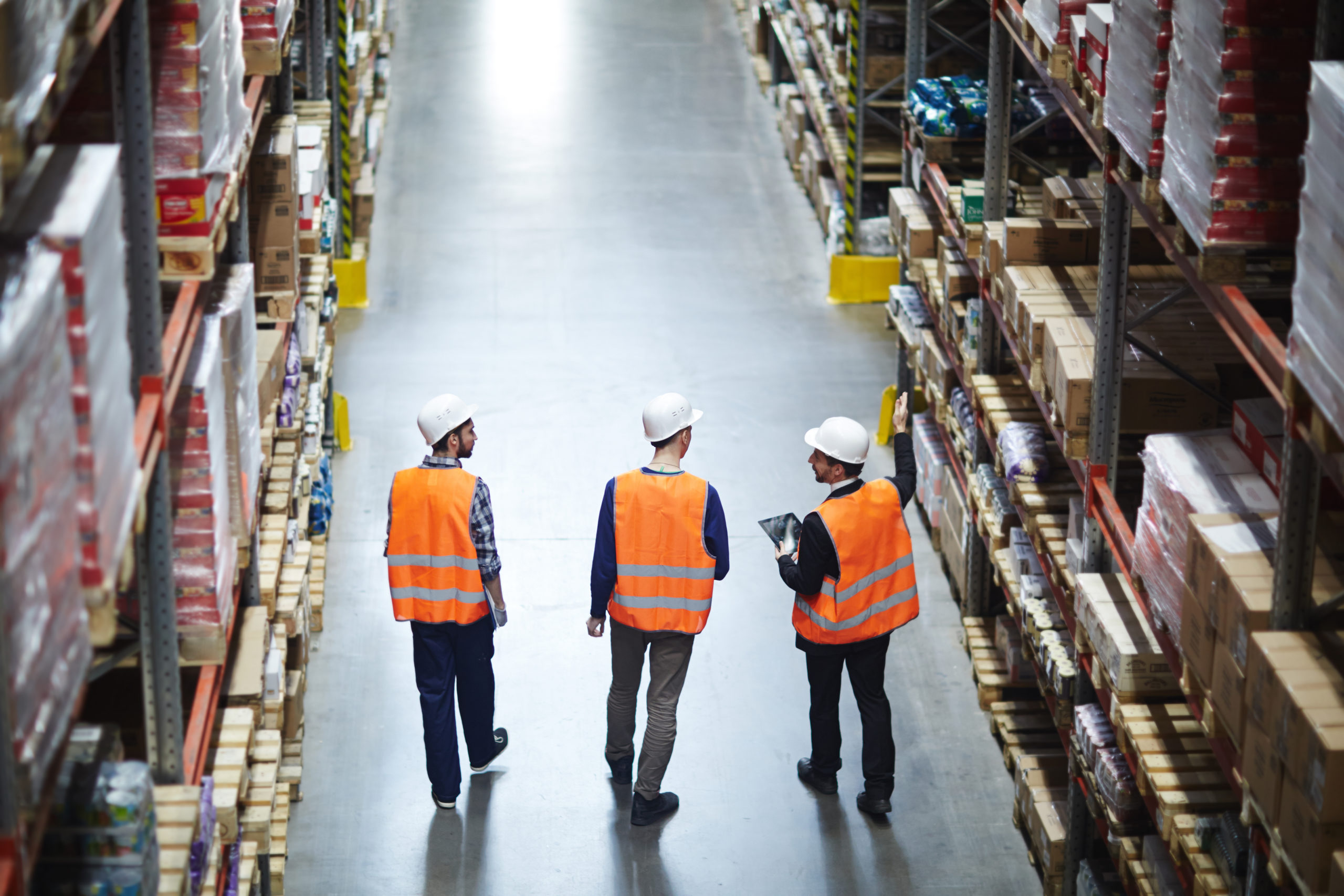 Group of warehouse workers wearing hardhats and reflective jackets waking in aisle between tall racks with packed goods, back view