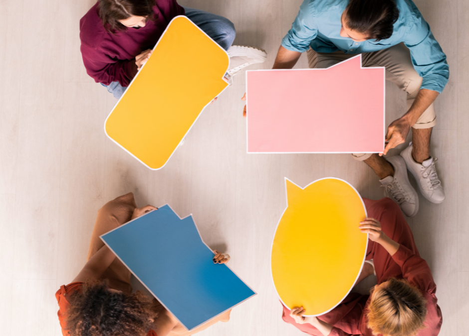 An overhead view of four people sitting on a light wooden floor, each holding a bright speech bubble in various colours (yellow, pink, blue, and orange). The image represents effective communication, teamwork, and stakeholder collaboration—key elements in return-to-work coordination and injury prevention strategies in Australian workplaces.