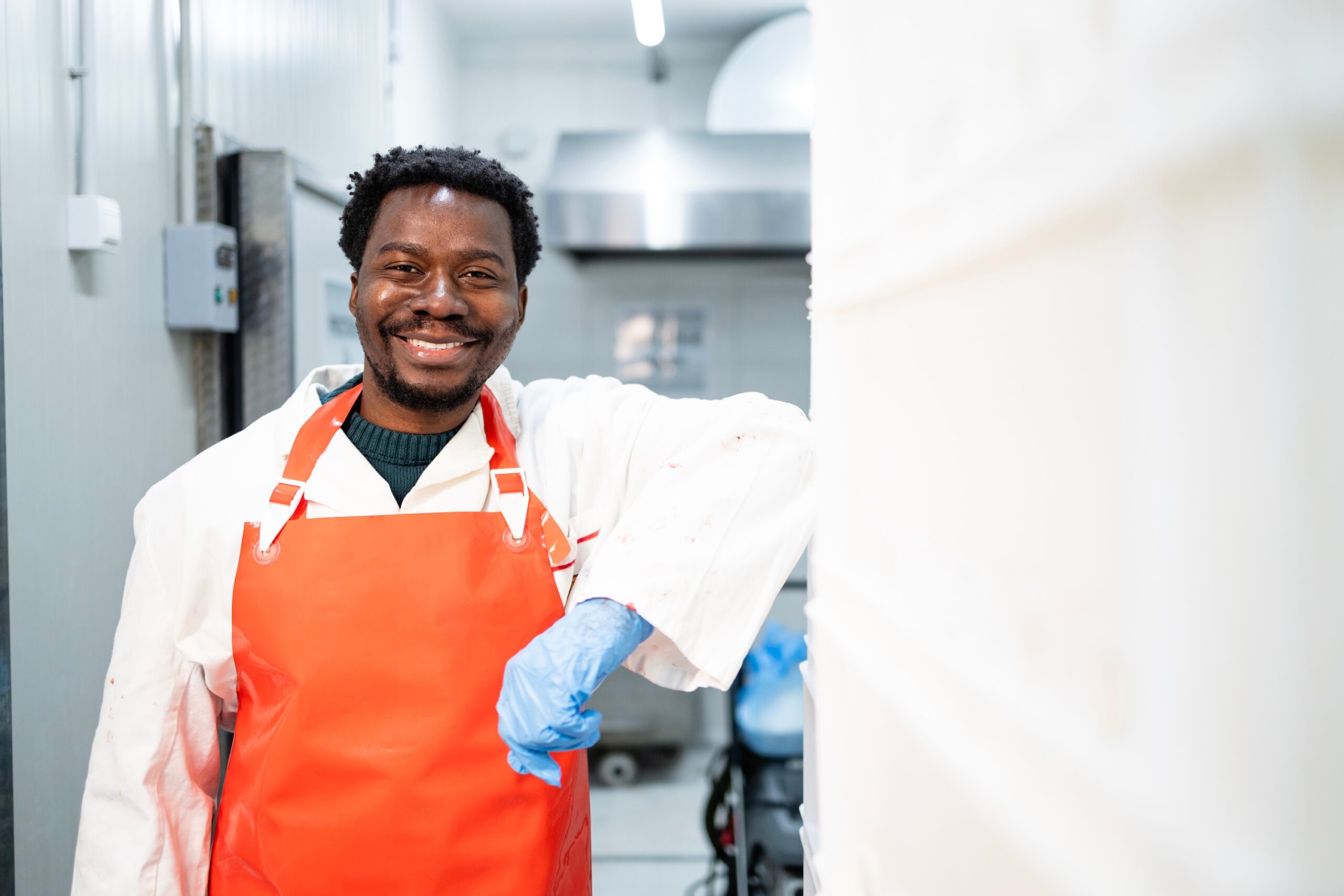 Smiling food production worker in a clean industrial setting, wearing a white coat, orange apron, and blue gloves, representing injury prevention and safe return-to-work practices.