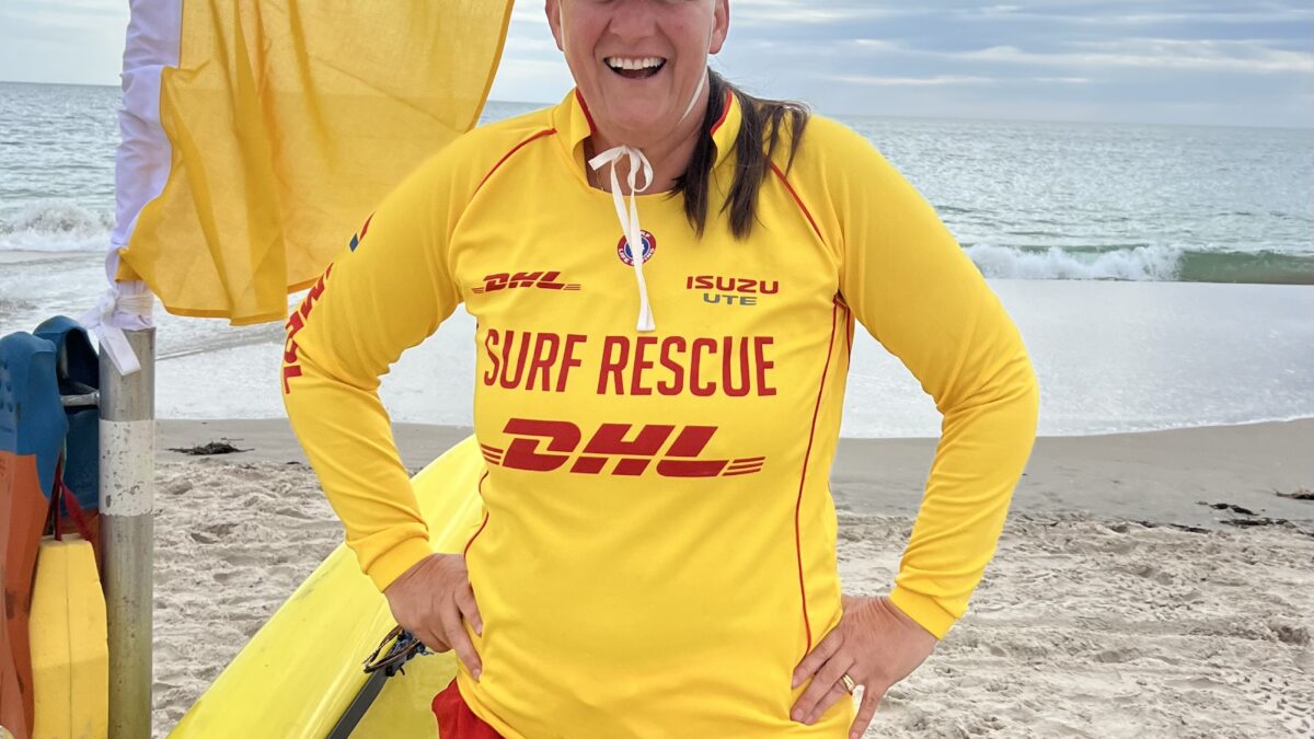 Image of Nicola Ross beside a surfboard and beach flags, wearing a surf life rescue shirt, ready to teach surf life rescue.