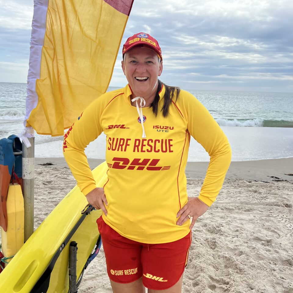 Image of Nicola Ross beside a surfboard and beach flags, wearing a surf life rescue shirt, ready to teach surf life rescue.
