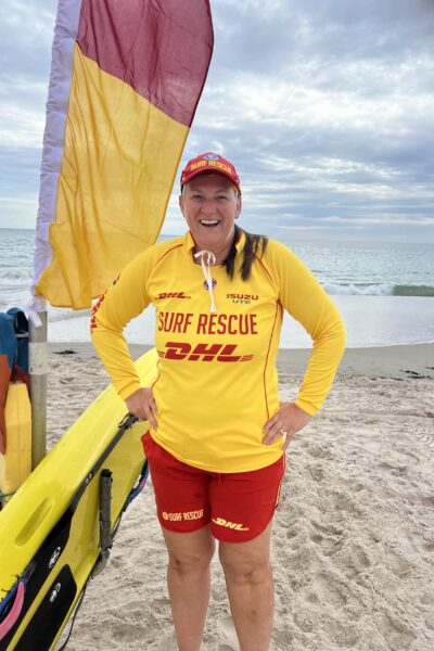 Image of Nicola Ross beside a surfboard and beach flags, wearing a surf life rescue shirt, ready to teach surf life rescue.