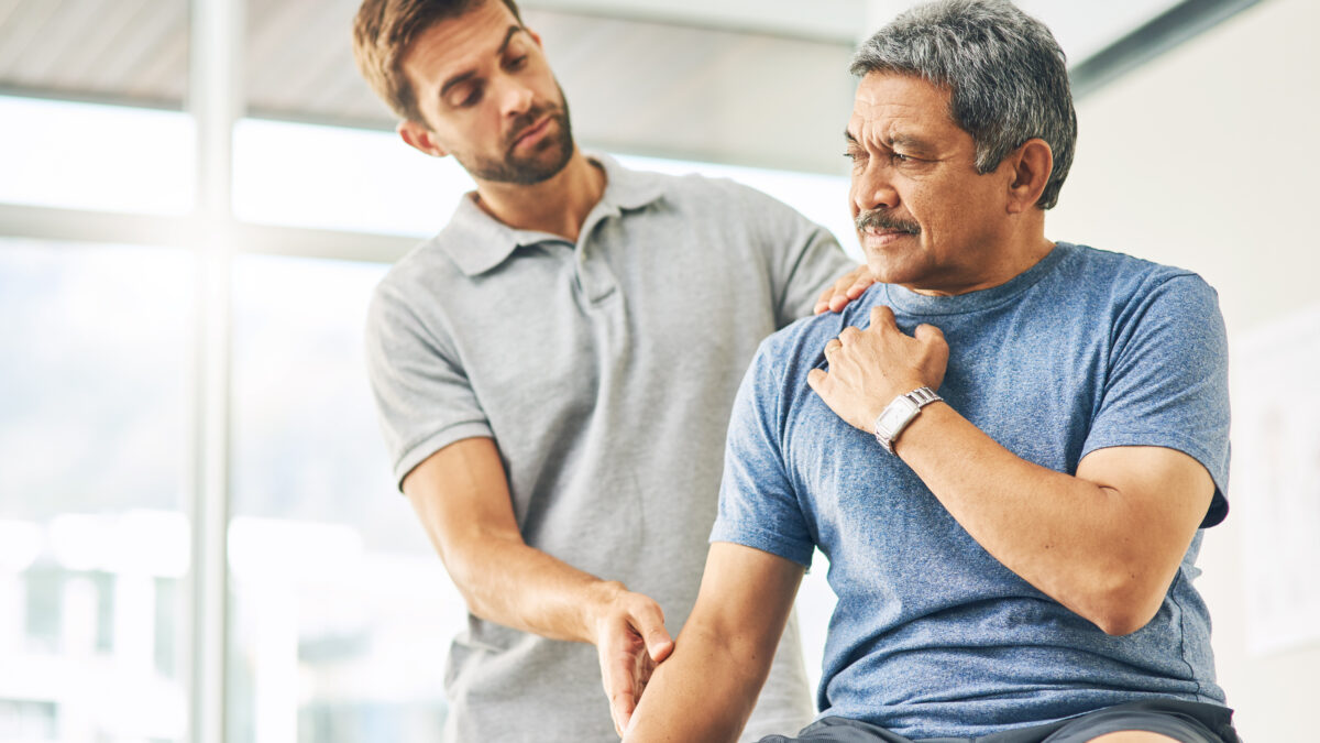 A physiotherapist is examining the shoulder and arm of a mature male worker wearing a blue t-shirt. The worker appears to be experiencing discomfort, suggesting early intervention for a musculoskeletal issue in a workplace setting. The scene is bright and professional, indicating an onsite health consultation.