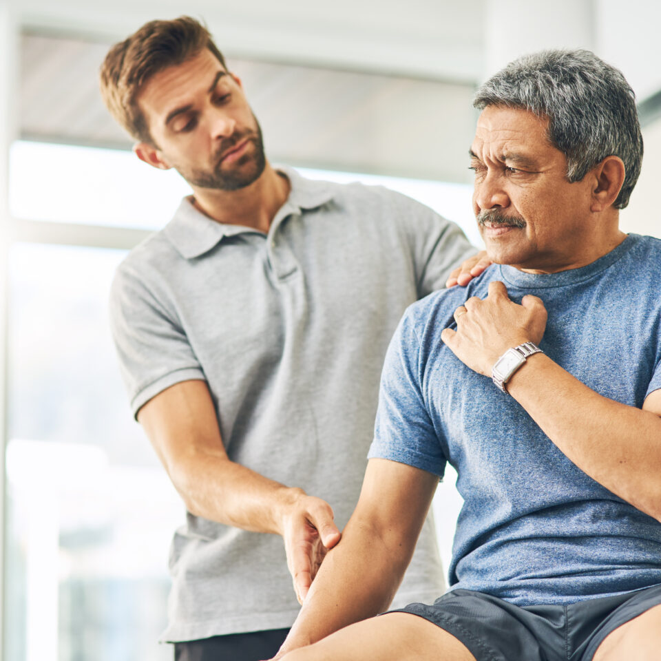 A physiotherapist is examining the shoulder and arm of a mature male worker wearing a blue t-shirt. The worker appears to be experiencing discomfort, suggesting early intervention for a musculoskeletal issue in a workplace setting. The scene is bright and professional, indicating an onsite health consultation.